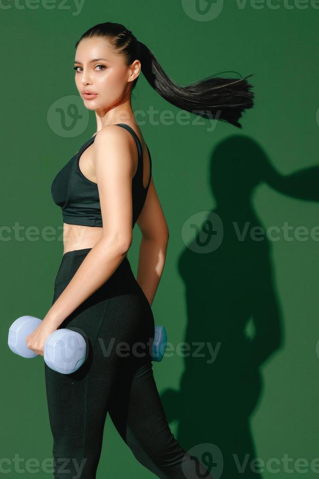 Beautiful strong cheerful young Asian sports woman does exercises with dumbbell isolated on green background. Fitness girl in sportswear posing indoors. Movement, Strength and motivation. photo