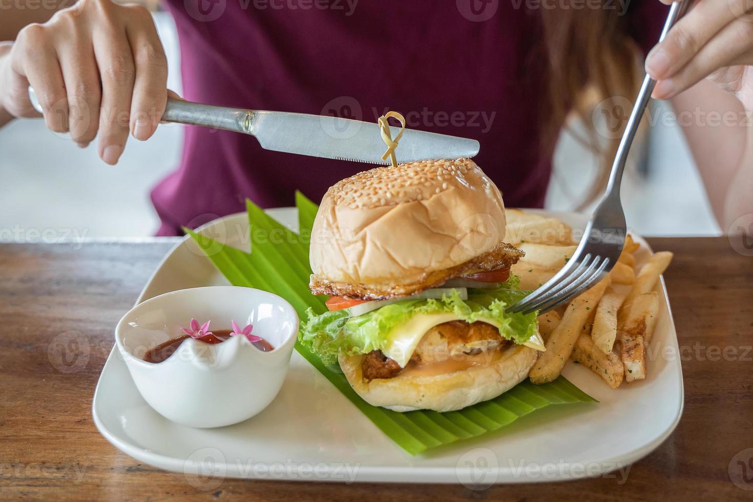 Knife and fork cutlery in hands with burger on banana leave with bowl of tomato sauce in white plate photo