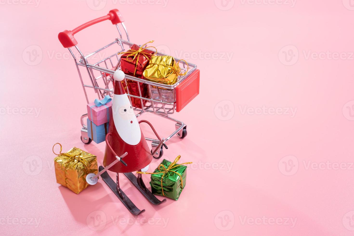 Christmas shopping concept, mini red shop cart trolley with Santa Claus toy and gift box isolated on pale pink background, blank copy space, close up photo