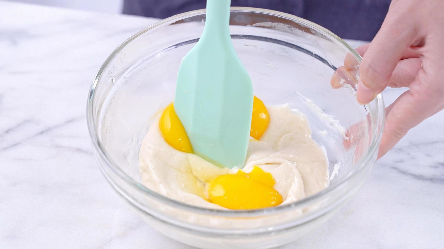 Mixing egg yolk into cake batter with green rubber spatula mixer tool stirring until smooth and blend well in a glass bowl, close up, lifestyle photo