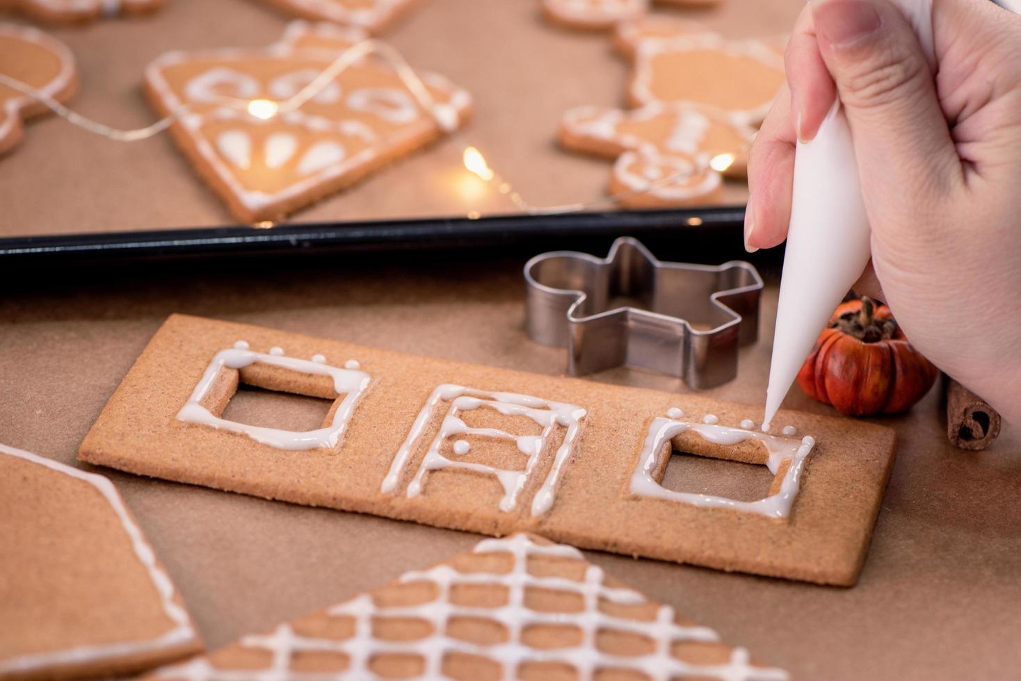 Woman is decorating gingerbread cookies house with white frosting icing cream topping on wooden table background, baking paper in kitchen, close up, macro. photo