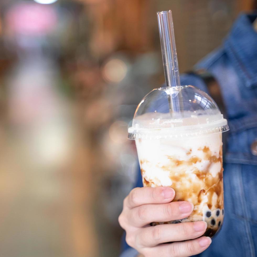 Young girl in denim jacket is drinking brown sugar flavored tapioca pearl bubble milk tea with glass straw in night market of Taiwan, close up, bokeh photo