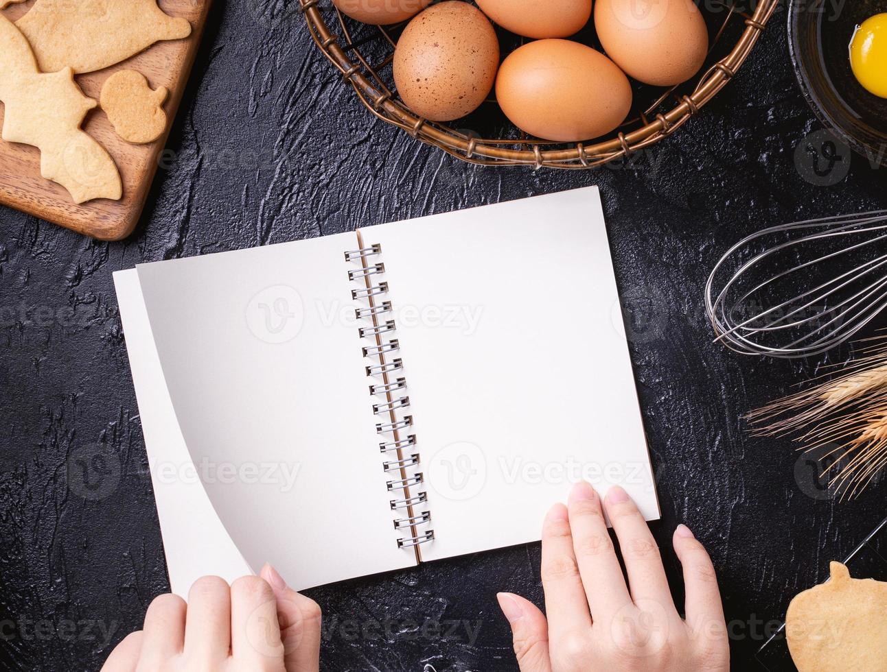 Woman is reading cookbook recipe of making Halloween cookies with baking ingredients, design concept of cooking class, top view, flat lay, overhead. photo