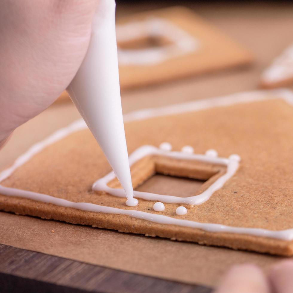 Woman is decorating gingerbread cookies house with white frosting icing cream topping on wooden table background, baking paper in kitchen, close up, macro. photo