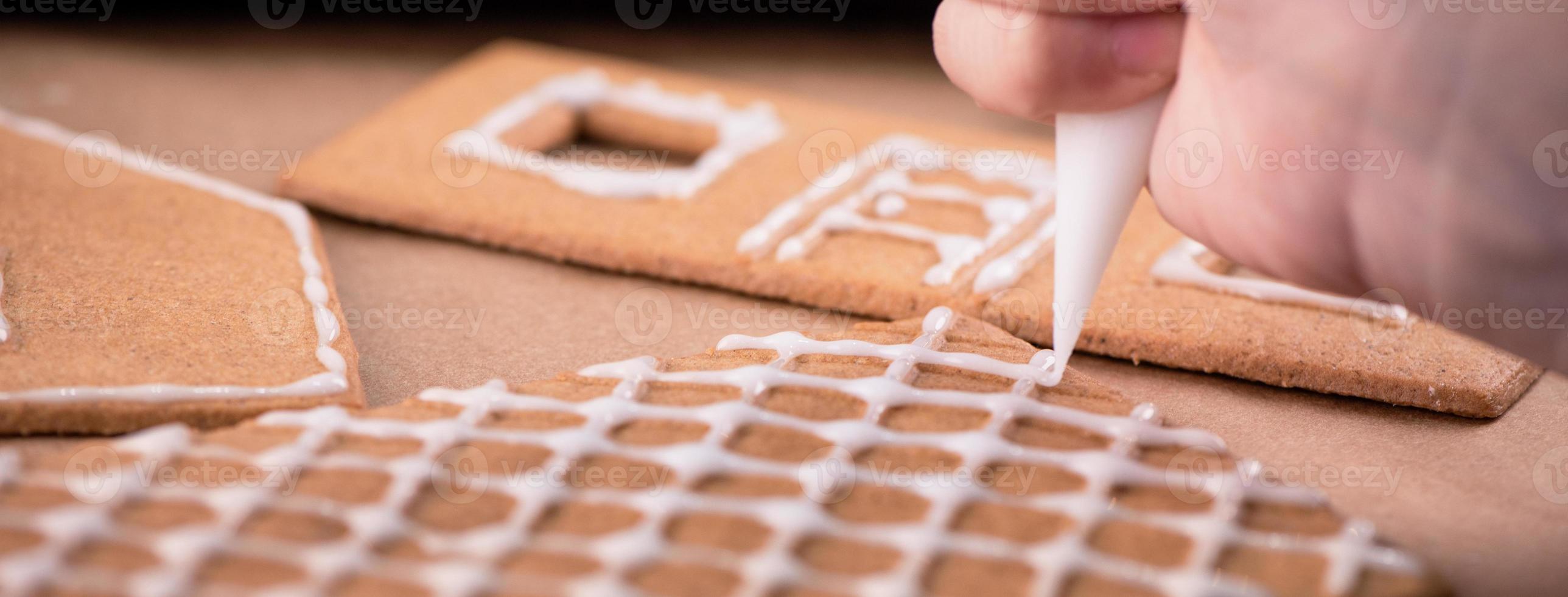 Woman is decorating gingerbread cookies house with white frosting icing cream topping on wooden table background, baking paper in kitchen, close up, macro. photo