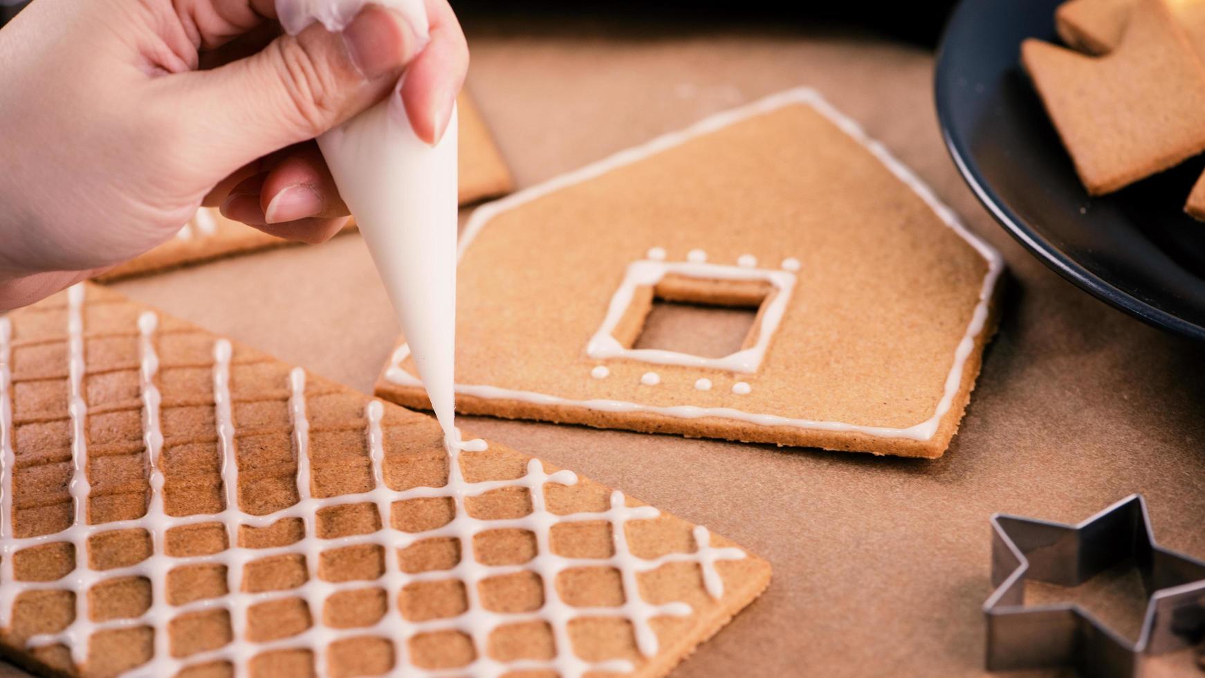 Woman is decorating gingerbread cookies house with white frosting icing cream topping on wooden table background, baking paper in kitchen, close up, macro. photo