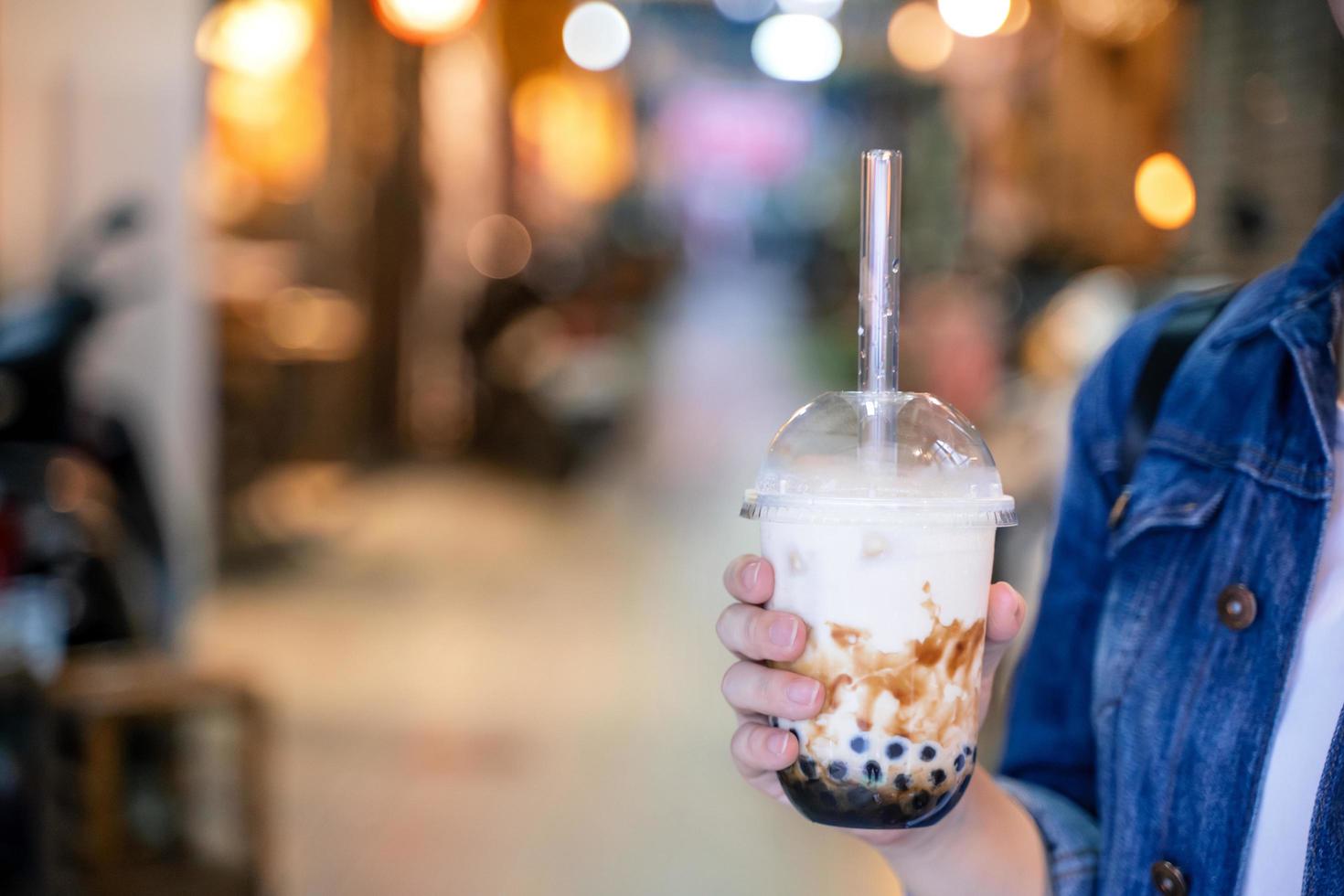 mujer joven está sosteniendo, bebiendo té de leche con burbujas de perlas de tapioca con sabor a azúcar moreno con paja de vidrio en el mercado nocturno de taiwán, de cerca, bokeh foto