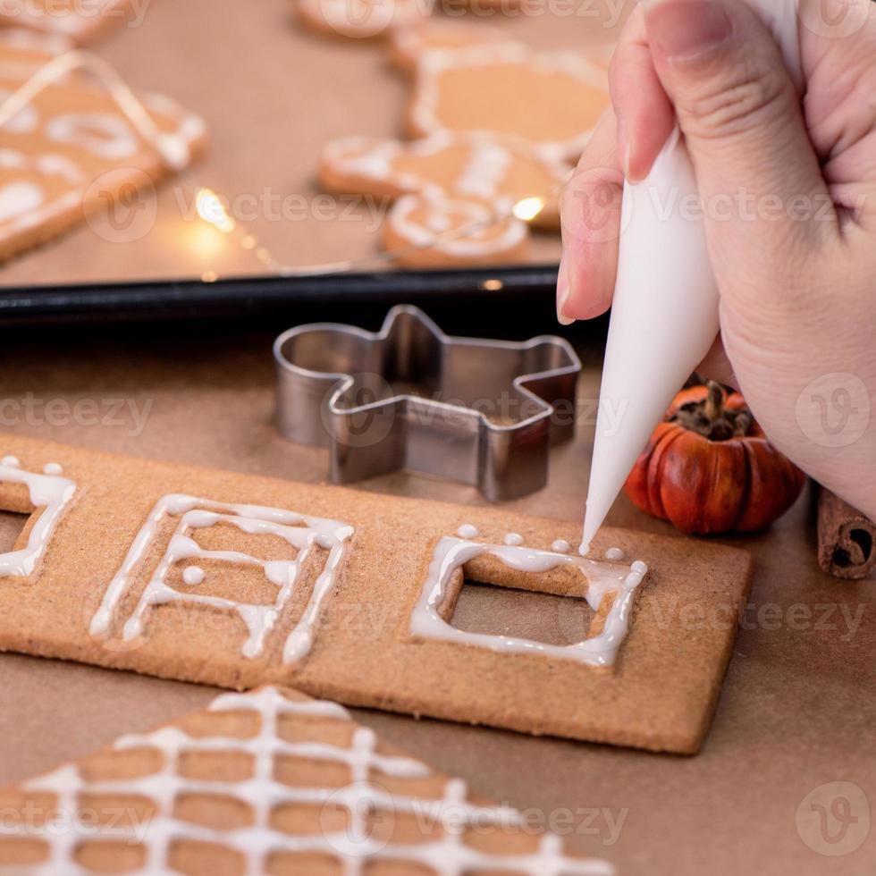 Woman is decorating gingerbread cookies house with white frosting icing cream topping on wooden table background, baking paper in kitchen, close up, macro. photo