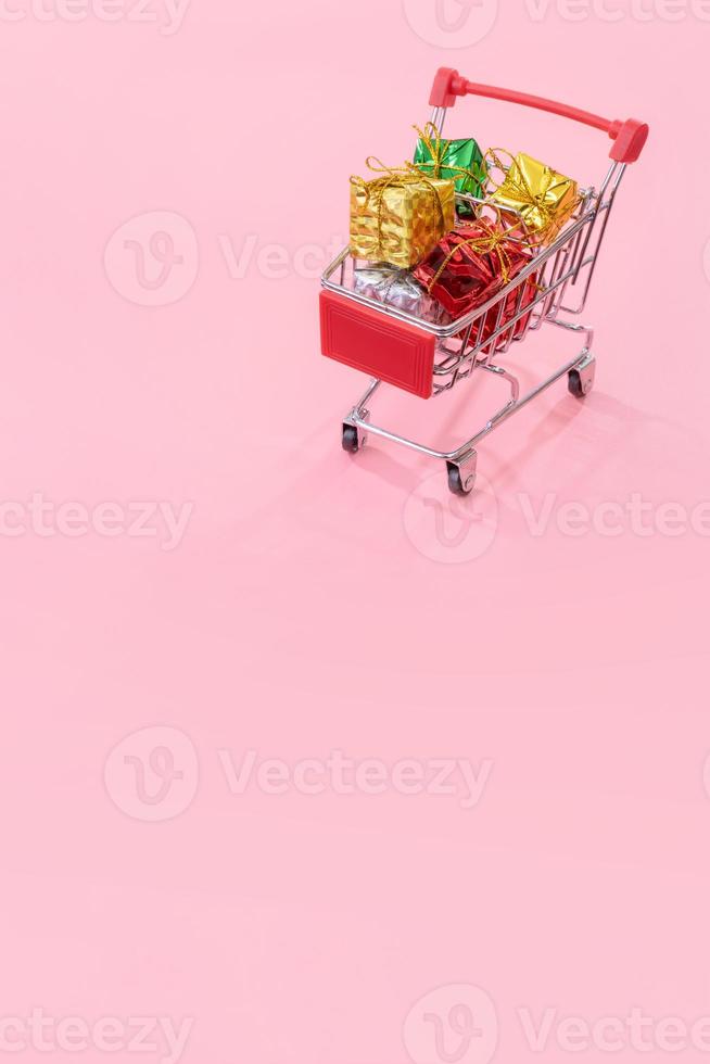 venta anual, concepto de temporada de compras navideñas - mini carrito de compras rojo lleno de caja de regalo aislado en fondo rosa pálido, espacio de copia, primer plano foto