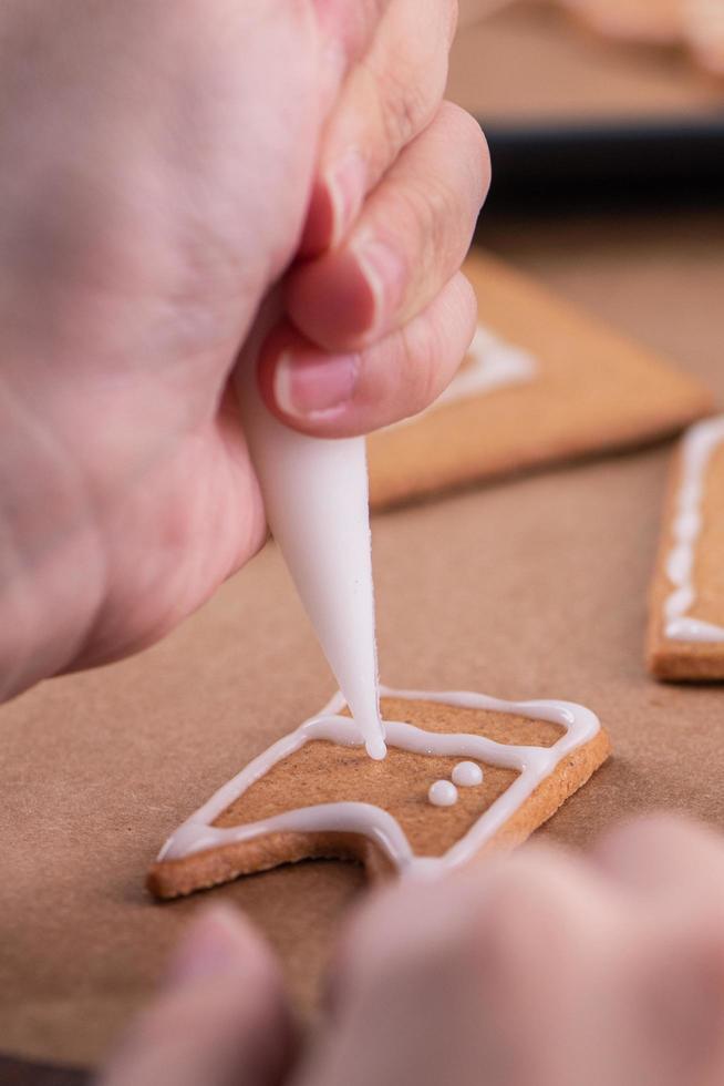 la mujer está decorando la casa de galletas de jengibre con cobertura de crema de hielo blanco sobre fondo de mesa de madera, papel para hornear en la cocina, primer plano, macro. foto