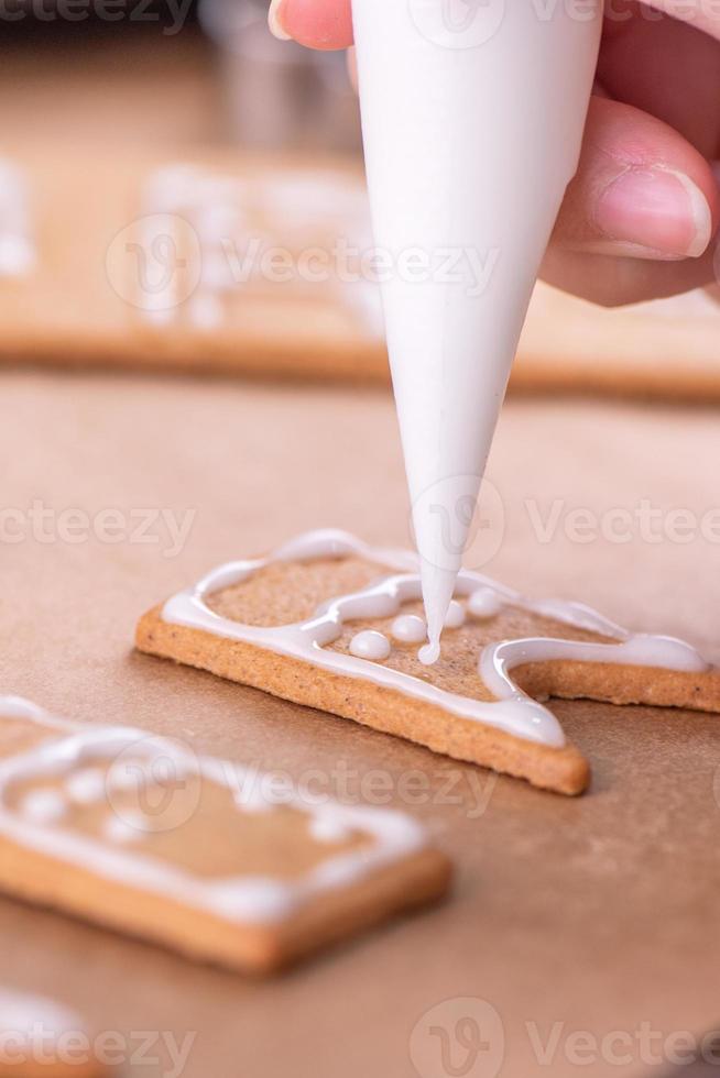 Woman is decorating gingerbread cookies house with white frosting icing cream topping on wooden table background, baking paper in kitchen, close up, macro. photo