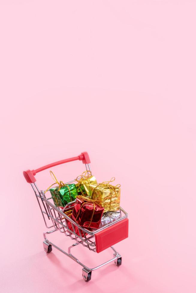 venta anual, concepto de temporada de compras navideñas - mini carrito de compras rojo lleno de caja de regalo aislado en fondo rosa pálido, espacio de copia, primer plano foto