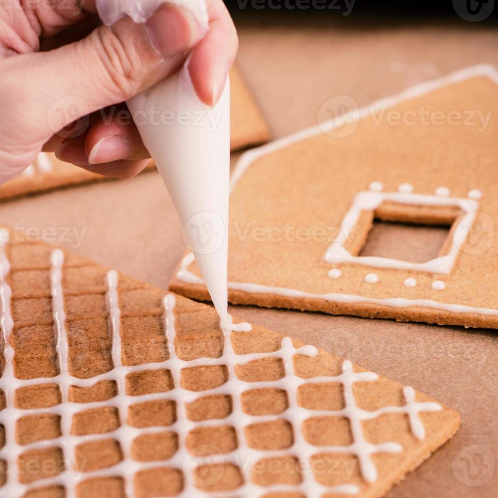 Woman is decorating gingerbread cookies house with white frosting icing cream topping on wooden table background, baking paper in kitchen, close up, macro. photo