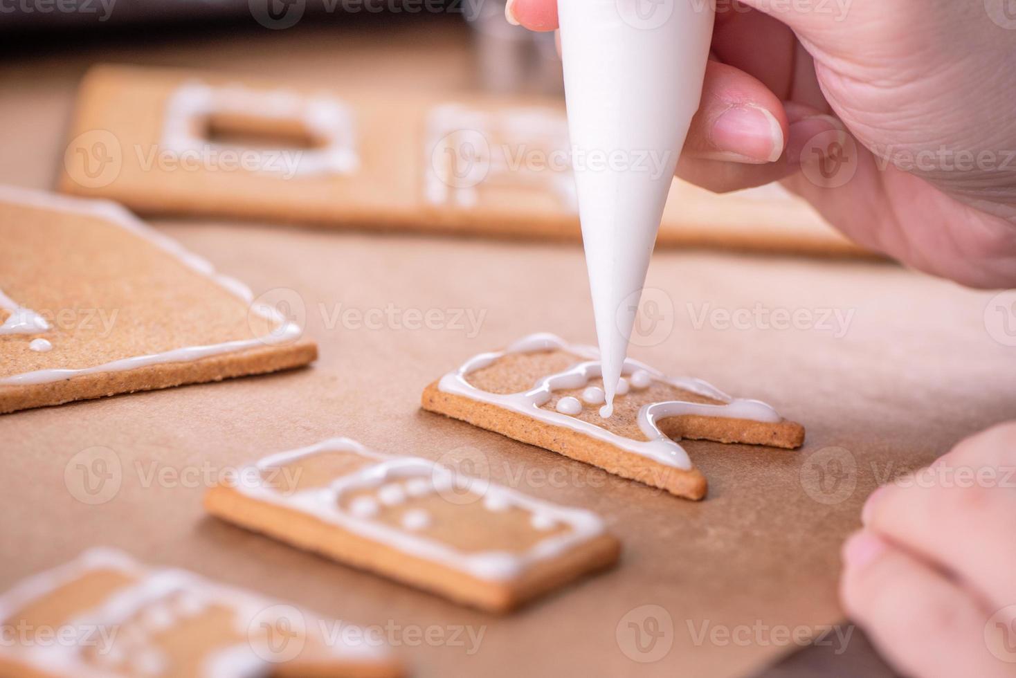 Woman is decorating gingerbread cookies house with white frosting icing cream topping on wooden table background, baking paper in kitchen, close up, macro. photo
