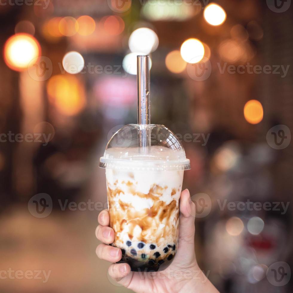 Young woman is holding, drinking brown sugar flavored tapioca pearl bubble milk tea with glass straw in night market of Taiwan, close up, bokeh photo