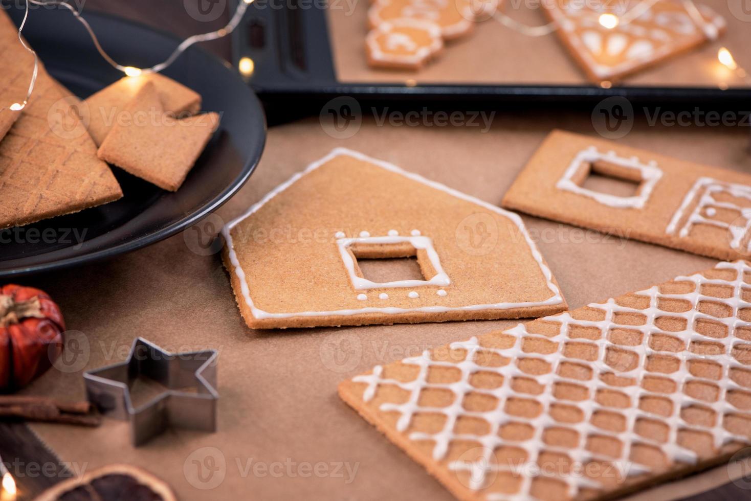 la mujer está decorando la casa de galletas de jengibre con cobertura de crema de hielo blanco sobre fondo de mesa de madera, papel para hornear en la cocina, primer plano, macro. foto