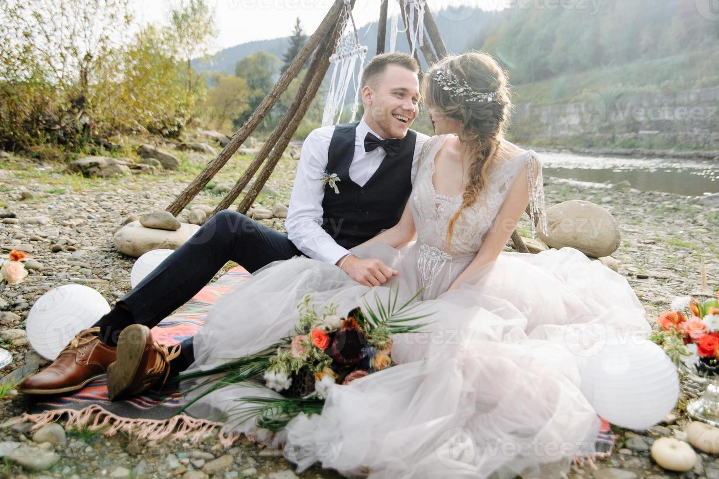 atractiva pareja de recién casados, momento feliz y alegre. el hombre y la mujer con ropa festiva se sientan en las piedras cerca de la decoración de la boda al estilo boho. ceremonia al aire libre. foto