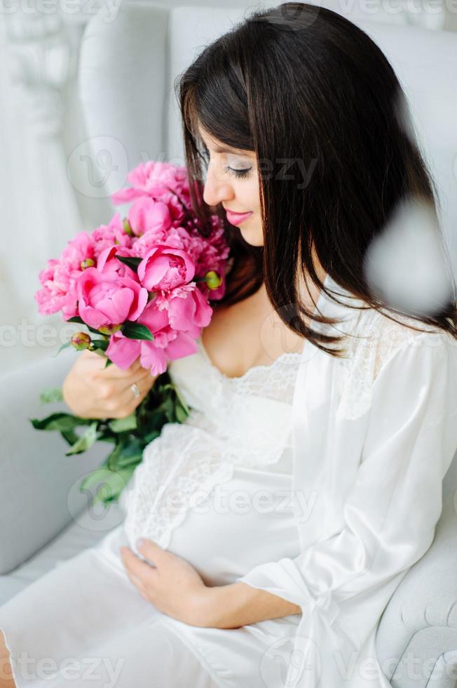 Close up photo of a pregnant woman with a naked belly holding a baby white booties and pink flowers on a white bed