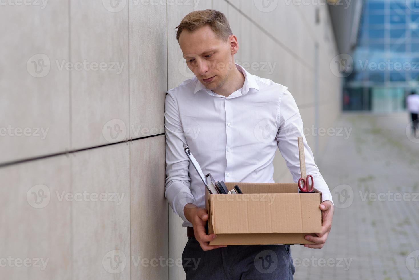 The fired office worker fell to his knees and covered his face due to stress. In front of him is a cardboard box with stationery. The man is unhappy due to a reduction in the robot. photo
