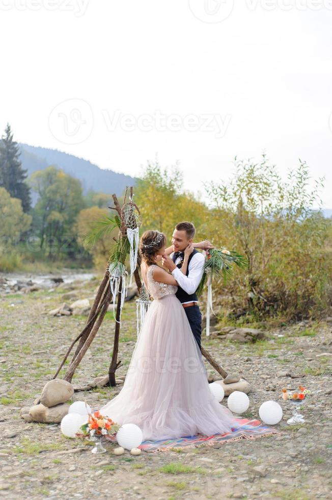 An attractive newlywed couple, a happy and joyful moment. A man and a woman shave and kiss in holiday clothes. Bohemian-style wedding cermonia in the forest in the fresh air. photo