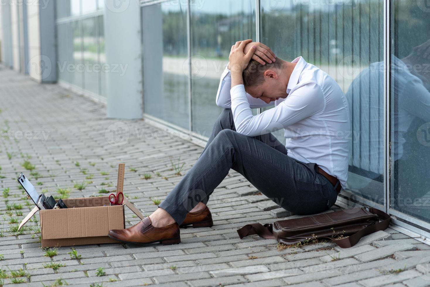 The fired office worker fell to his knees and covered his face due to stress. In front of him is a cardboard box with stationery. The man is unhappy due to a reduction in the robot. photo