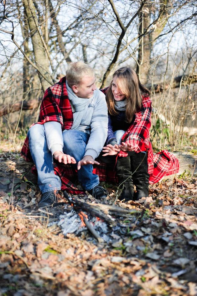 Young loving couple dressed in blue sweater sitting with red coffee cups on the firewood in winter forest photo