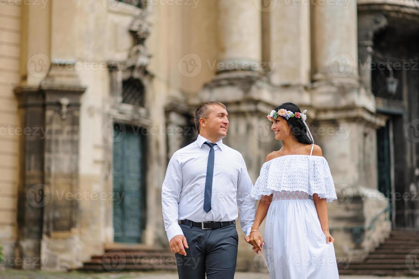 Wedding photo session on the background of the old building. The groom watches his bride posing. Rustic or boho wedding photography.