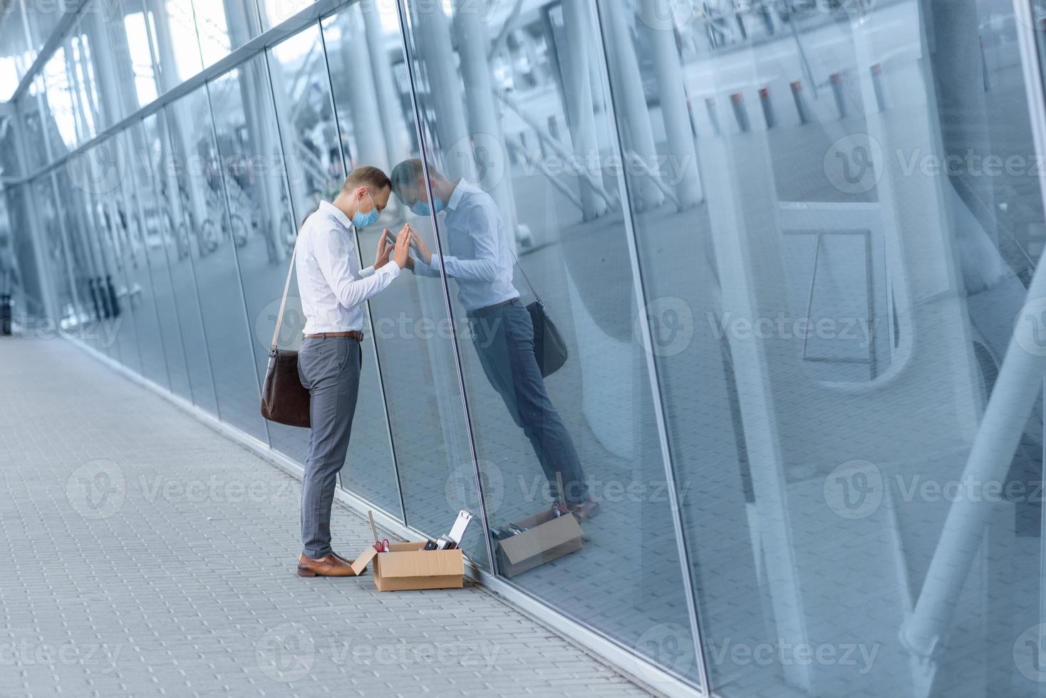 The fired office worker fell to his knees and covered his face due to stress. In front of him is a cardboard box with stationery. The man is unhappy due to a reduction in the robot. photo