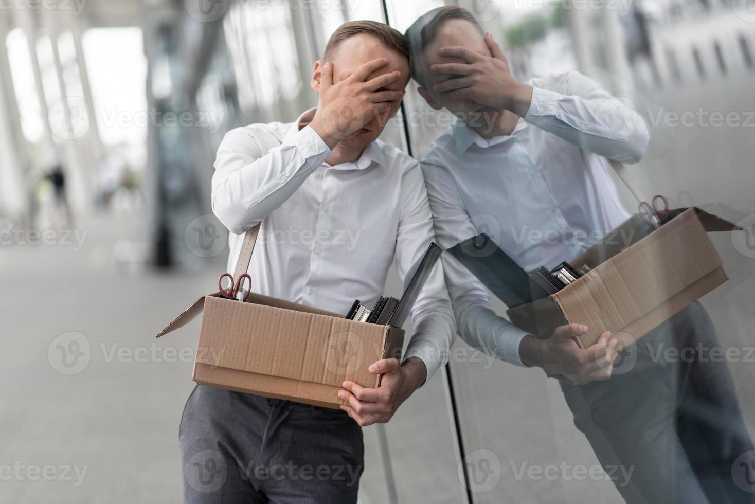 The fired office worker fell to his knees and covered his face due to stress. In front of him is a cardboard box with stationery. The man is unhappy due to a reduction in the robot. photo