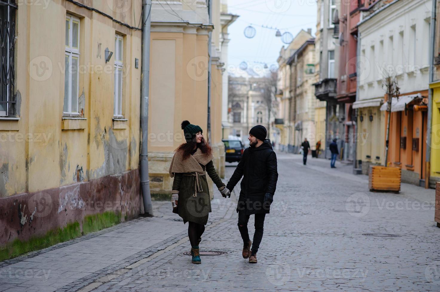 una pareja joven de moda camina por la ciudad, la joven tiene bolsas de compras en el brazo y el hombre una taza de café en la mano foto