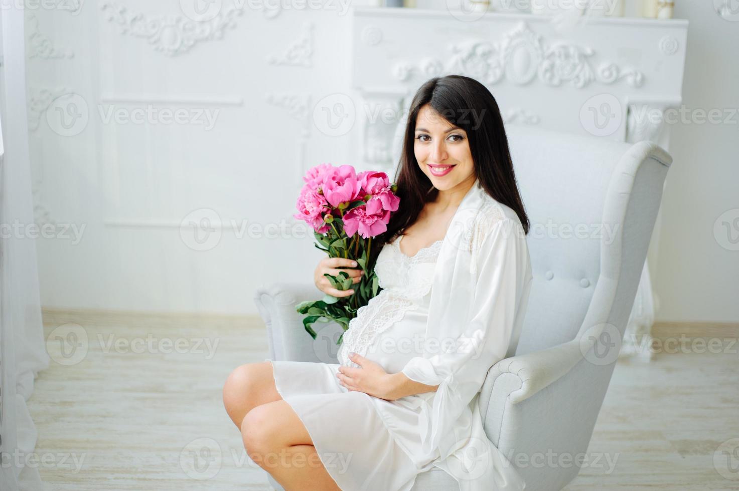 Close up photo of a pregnant woman with a naked belly holding a baby white booties and pink flowers on a white bed