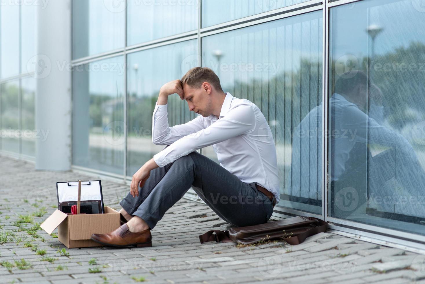 The fired office worker fell to his knees and covered his face due to stress. In front of him is a cardboard box with stationery. The man is unhappy due to a reduction in the robot. photo