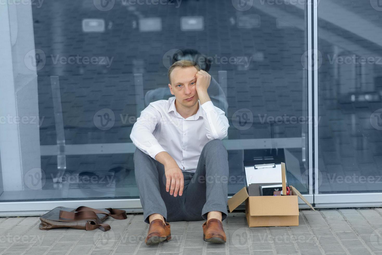 The fired office worker fell to his knees and covered his face due to stress. In front of him is a cardboard box with stationery. The man is unhappy due to a reduction in the robot. photo