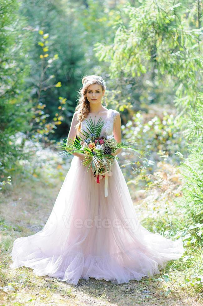 Happy bride in a pink wedding dress. The girl holds a wedding bouquet in her hands. Boho style wedding ceremony in the forest. photo