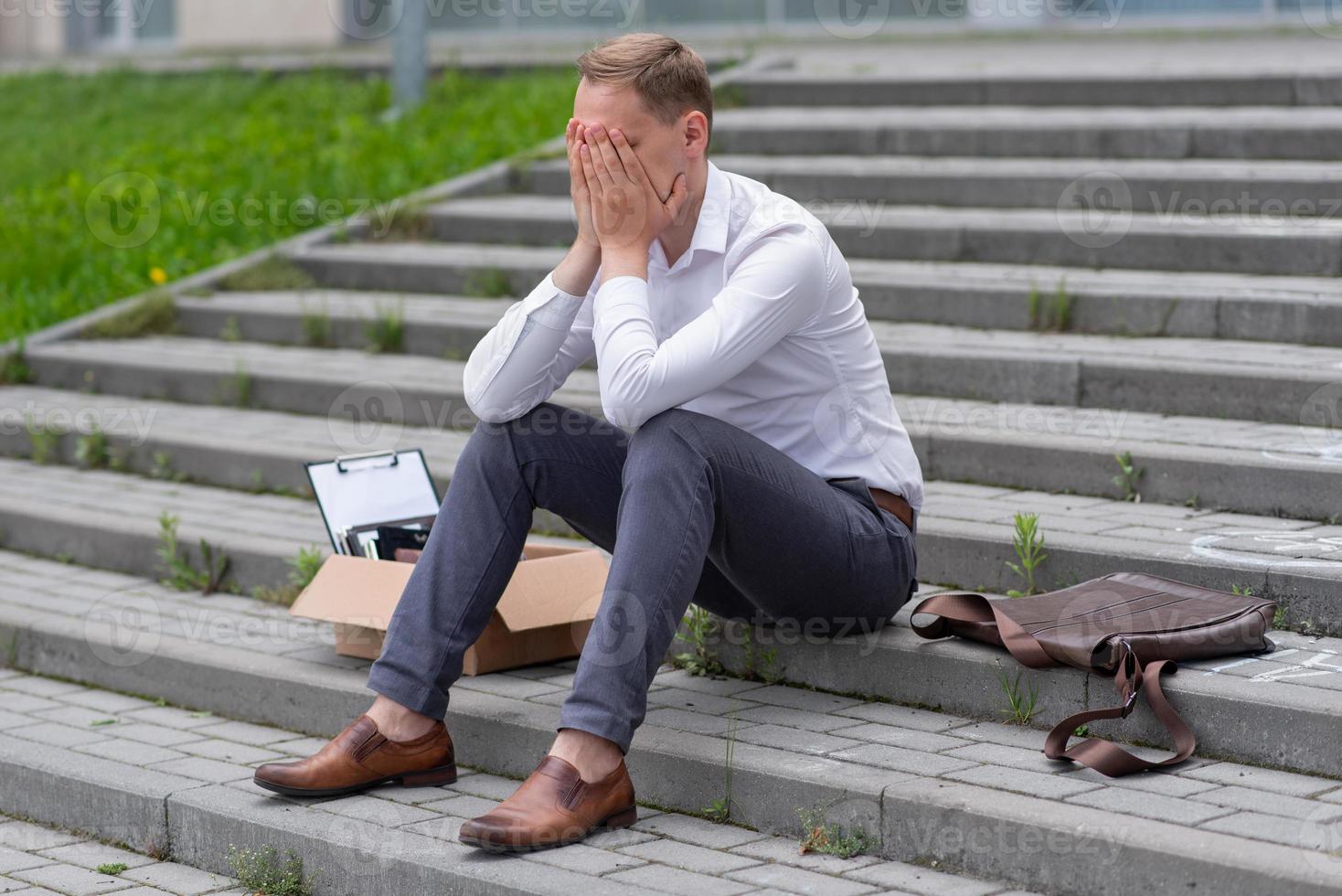 The fired office worker fell to his knees and covered his face due to stress. In front of him is a cardboard box with stationery. The man is unhappy due to a reduction in the robot. photo