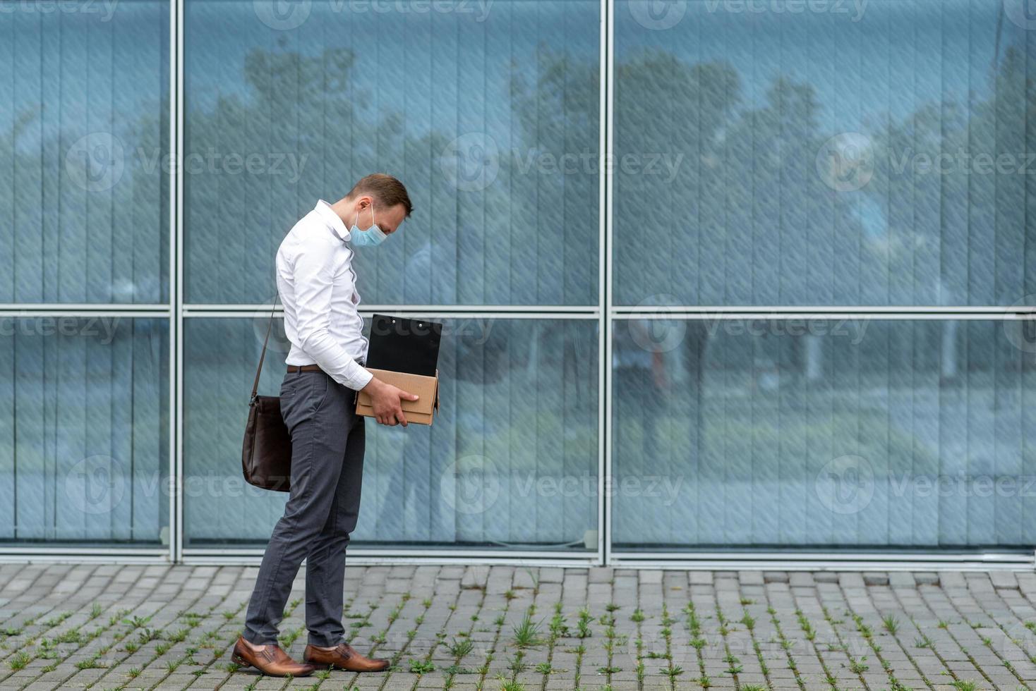 The fired office worker fell to his knees and covered his face due to stress. In front of him is a cardboard box with stationery. The man is unhappy due to a reduction in the robot. photo