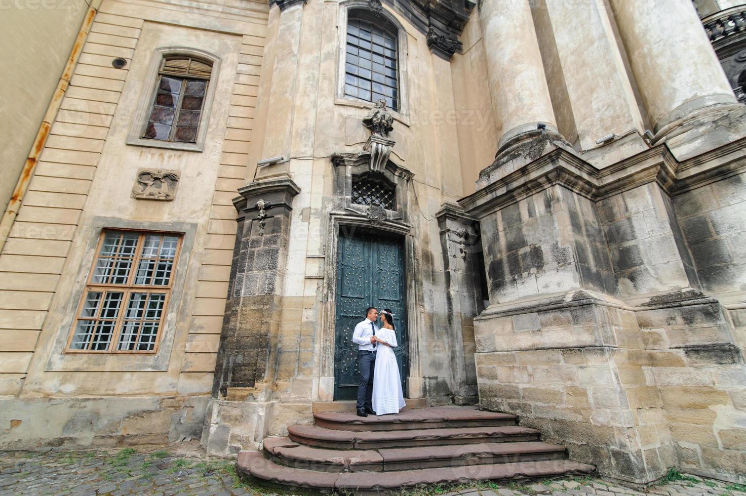 Wedding photo session on the background of the old building. The groom watches his bride posing. Rustic or boho wedding photography.