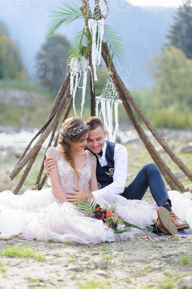 una atractiva pareja de recién casados, un momento feliz y alegre. un hombre y una mujer se afeitan y se besan con ropa de fiesta. ceremonia de boda de estilo bohemio en el bosque al aire libre. foto
