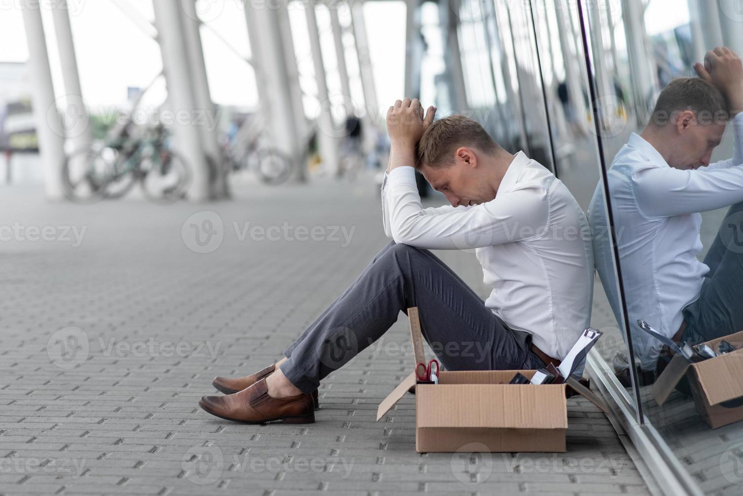 The fired office worker fell to his knees and covered his face due to stress. In front of him is a cardboard box with stationery. The man is unhappy due to a reduction in the robot. photo