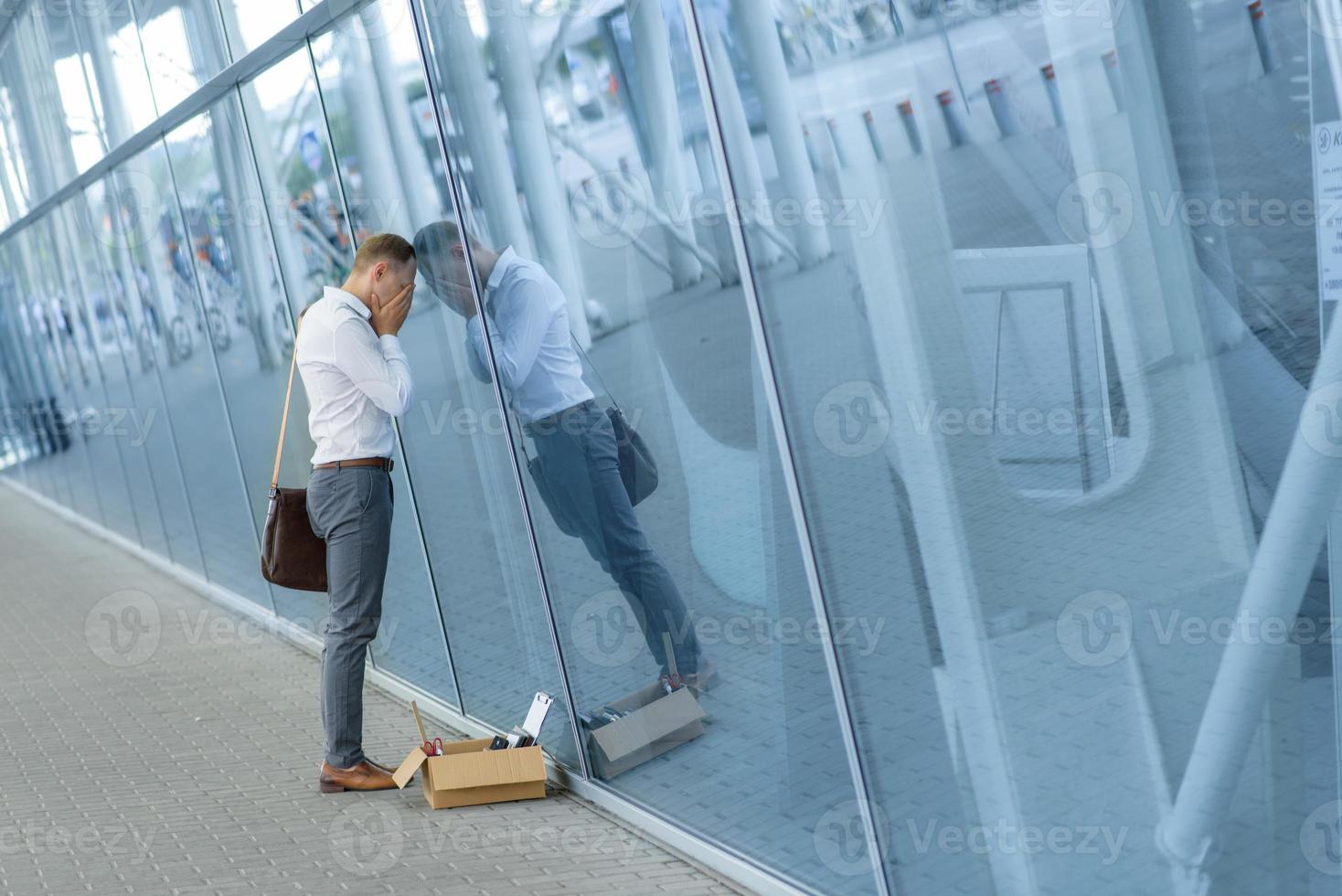 The fired office worker fell to his knees and covered his face due to stress. In front of him is a cardboard box with stationery. The man is unhappy due to a reduction in the robot. photo