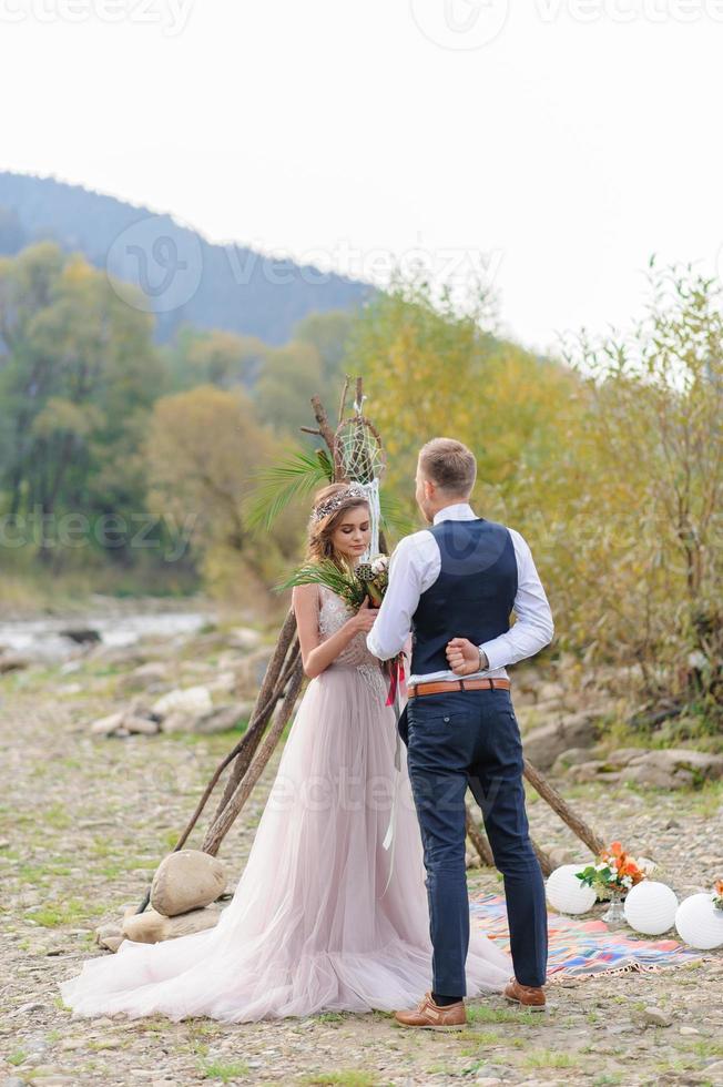 una atractiva pareja de recién casados, un momento feliz y alegre. un hombre y una mujer se afeitan y se besan con ropa de fiesta. ceremonia de boda de estilo bohemio en el bosque al aire libre. foto