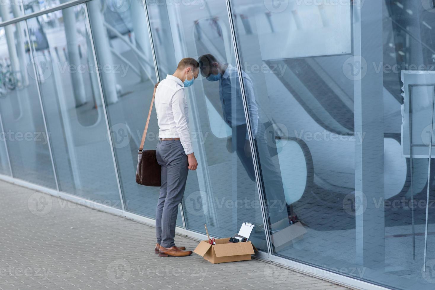 The fired office worker fell to his knees and covered his face due to stress. In front of him is a cardboard box with stationery. The man is unhappy due to a reduction in the robot. photo