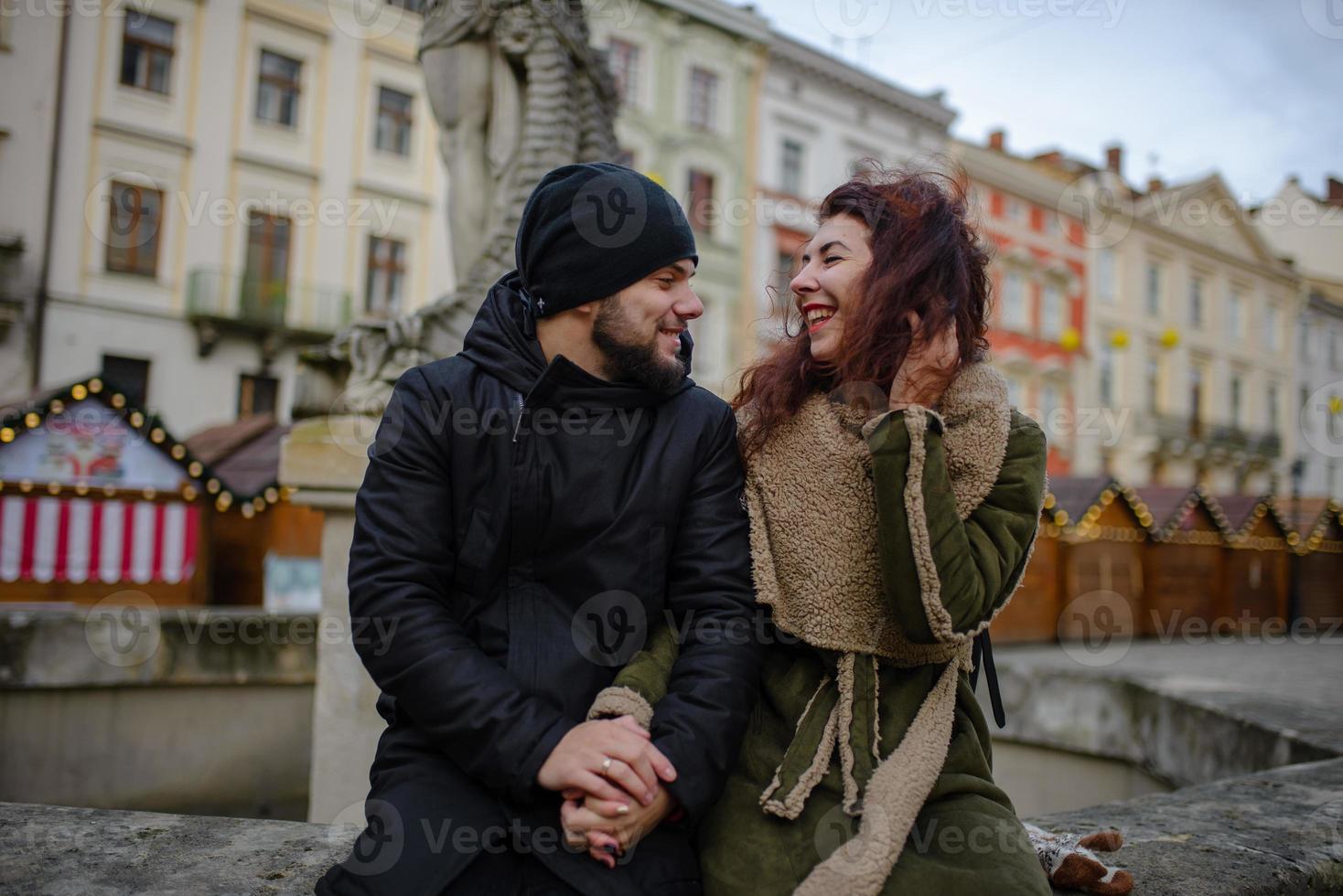 A young romantic couple wearing warm clothes outdoor at Christmas time, sitting on a bench in evening street decorated with beautiful lights. photo