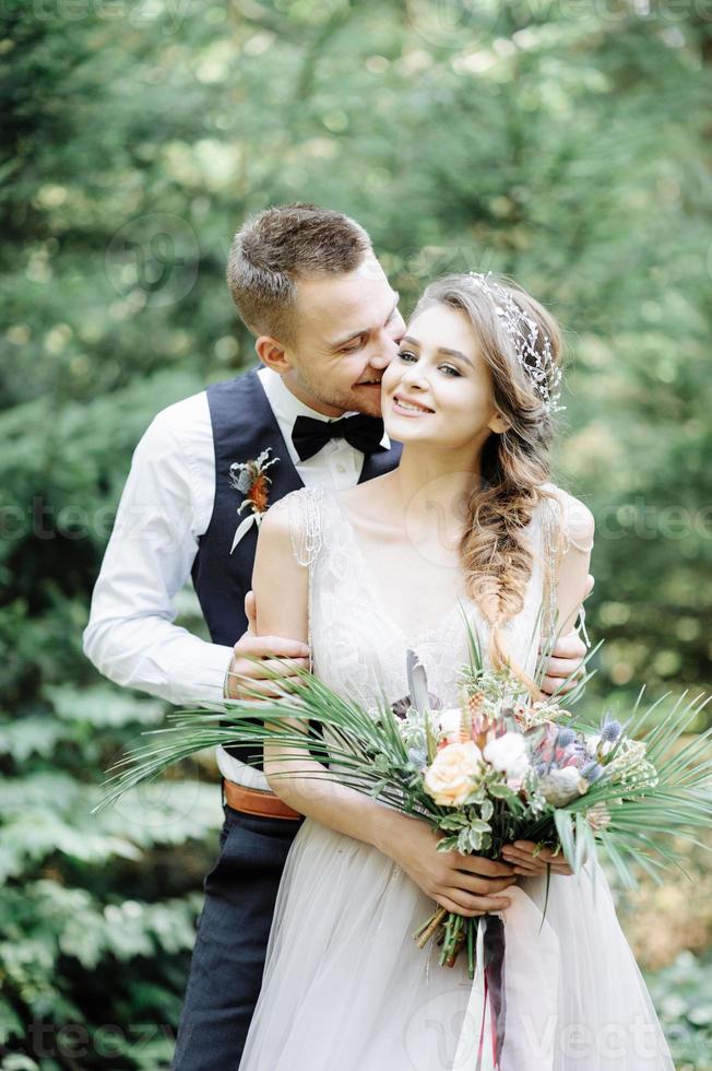 Attractive couple newlyweds, happy and joyful moment. Man and woman in festive clothes sit on the stones near the wedding decoration in boho style. Ceremony outdoors. photo