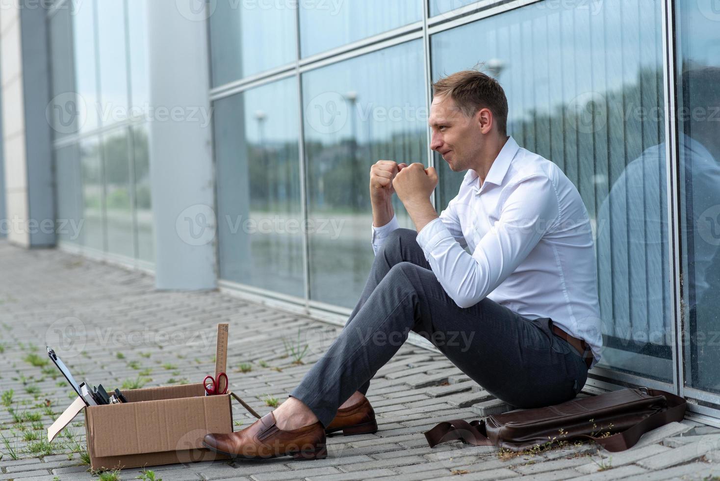 The fired office worker fell to his knees and covered his face due to stress. In front of him is a cardboard box with stationery. The man is unhappy due to a reduction in the robot. photo