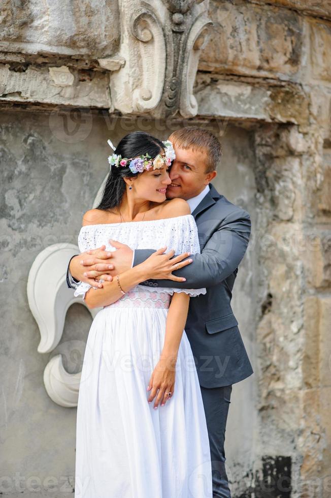 Wedding photo session on the background of the old building. The groom watches his bride posing. Rustic or boho wedding photography.