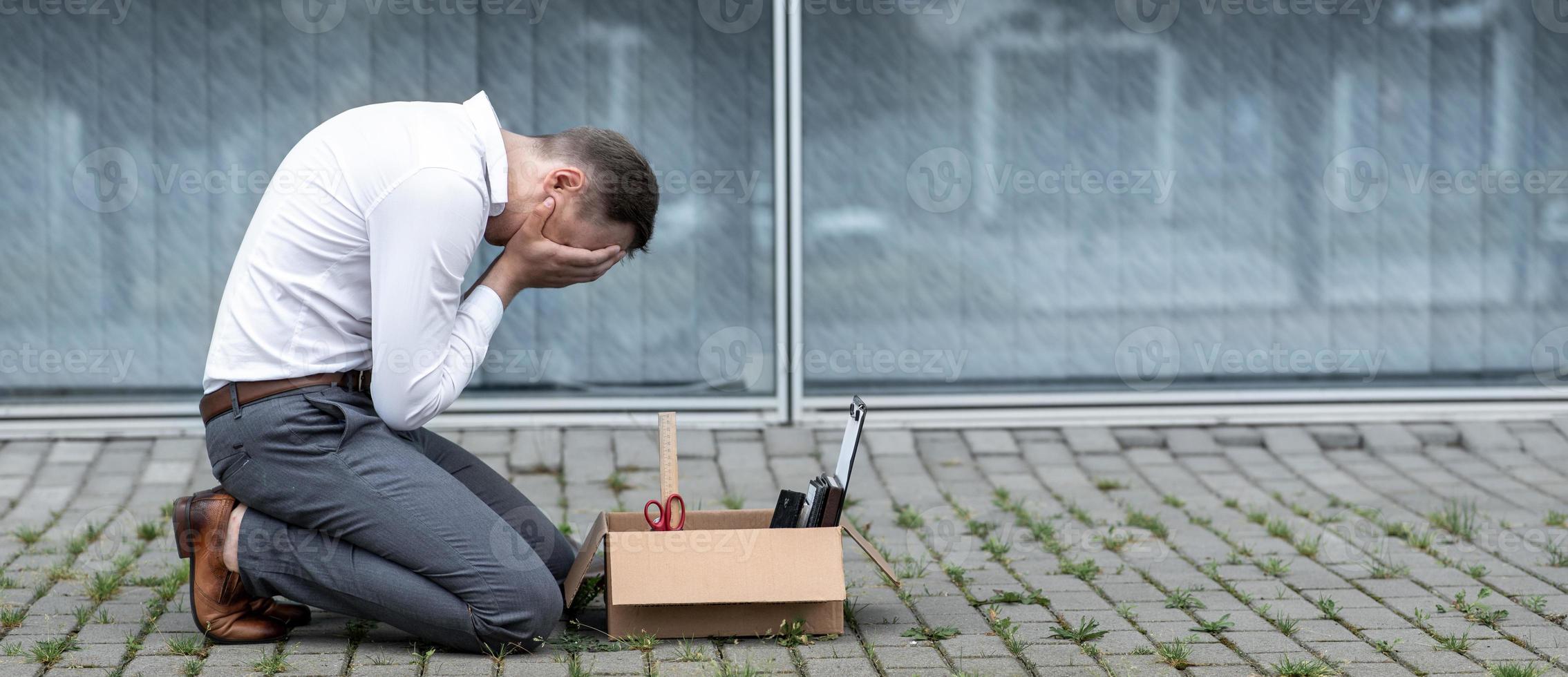 The fired office worker fell to his knees and covered his face due to stress. In front of him is a cardboard box with stationery. The man is unhappy due to a reduction in the robot. photo