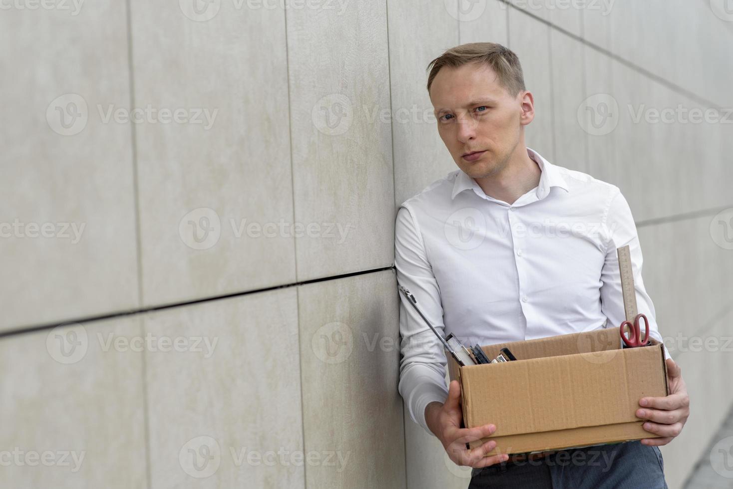The fired office worker fell to his knees and covered his face due to stress. In front of him is a cardboard box with stationery. The man is unhappy due to a reduction in the robot. photo