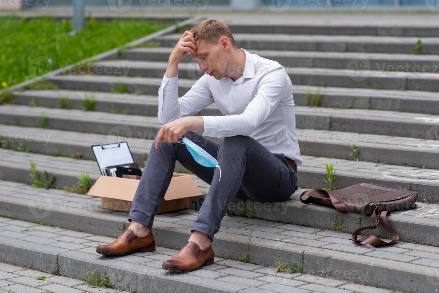 The fired office worker fell to his knees and covered his face due to stress. In front of him is a cardboard box with stationery. The man is unhappy due to a reduction in the robot. photo