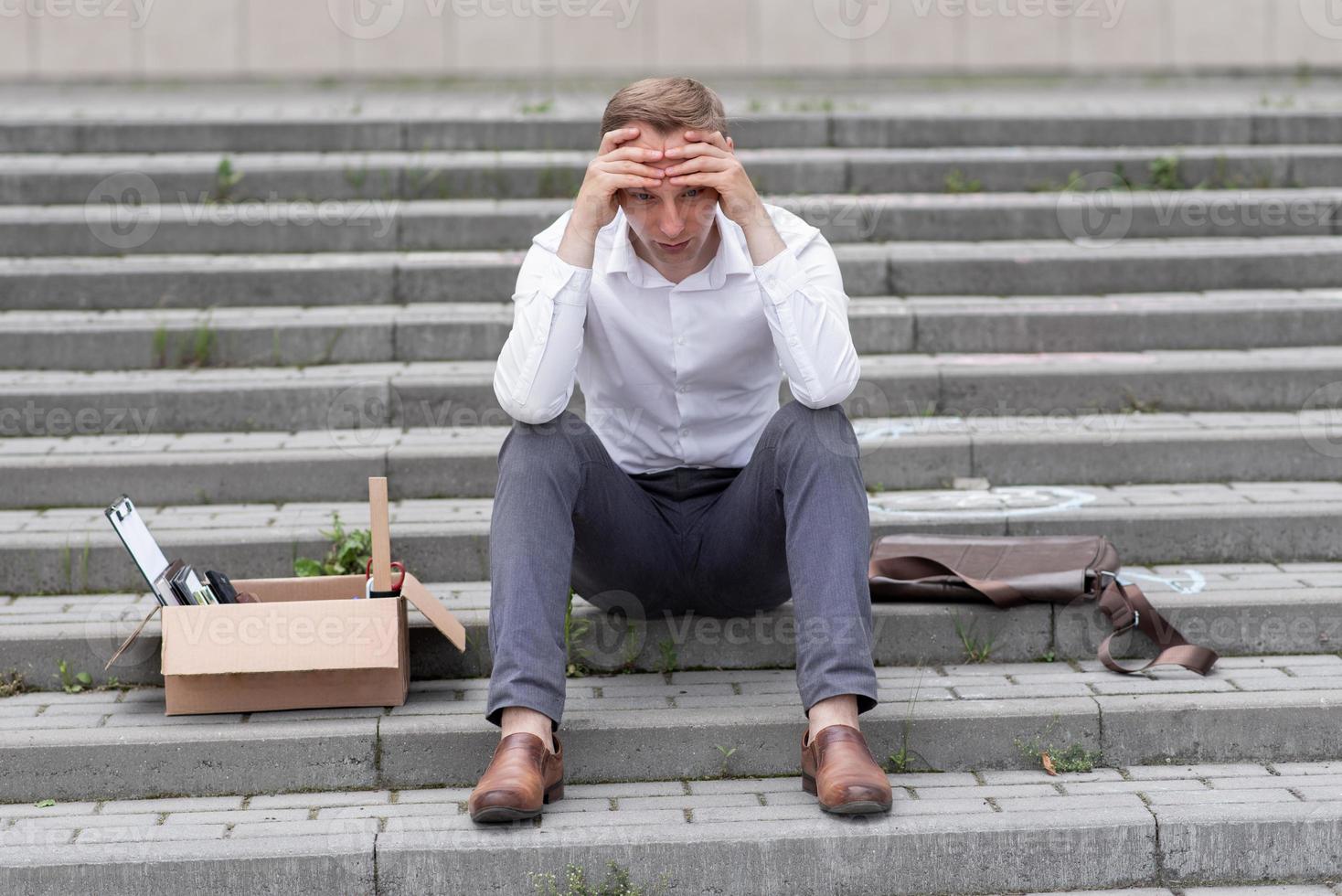 The fired office worker fell to his knees and covered his face due to stress. In front of him is a cardboard box with stationery. The man is unhappy due to a reduction in the robot. photo
